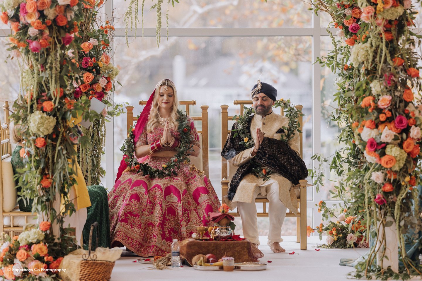Bride and groom in traditional attire sit under colorful floral arch.