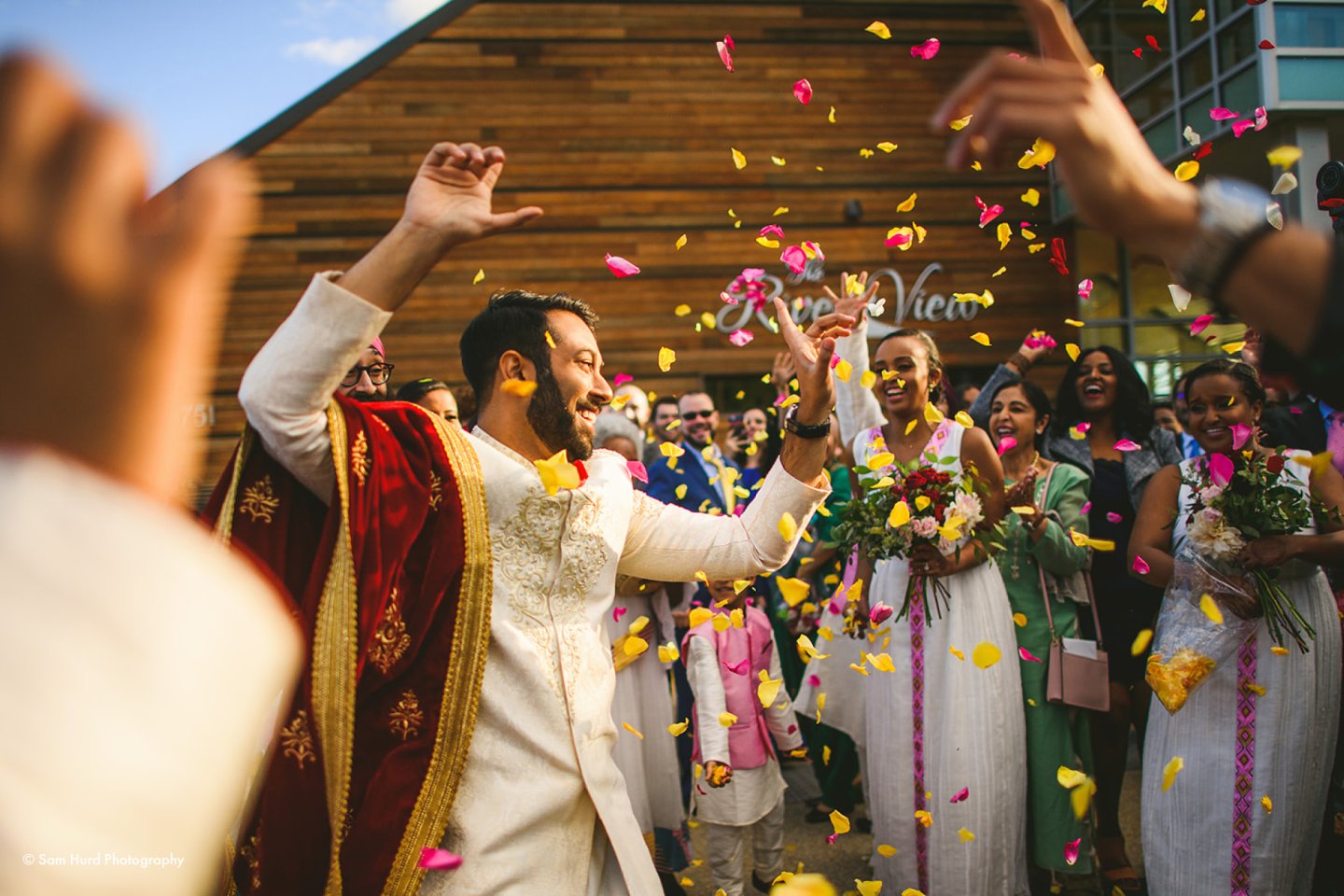 Man celebrating with friends, colorful petals in the air, vibrant wedding scene.