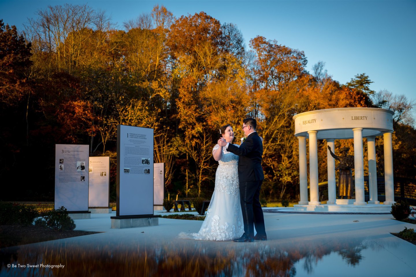 Wedding couple dancing by a white pavilion, surrounded by autumn trees.