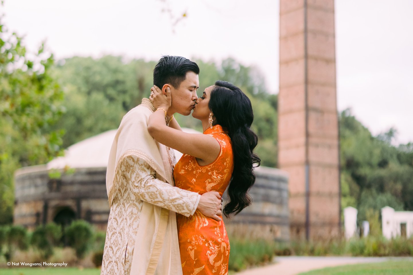 Couple kissing outdoors, wearing traditional attire, with greenery and a brick tower behind.