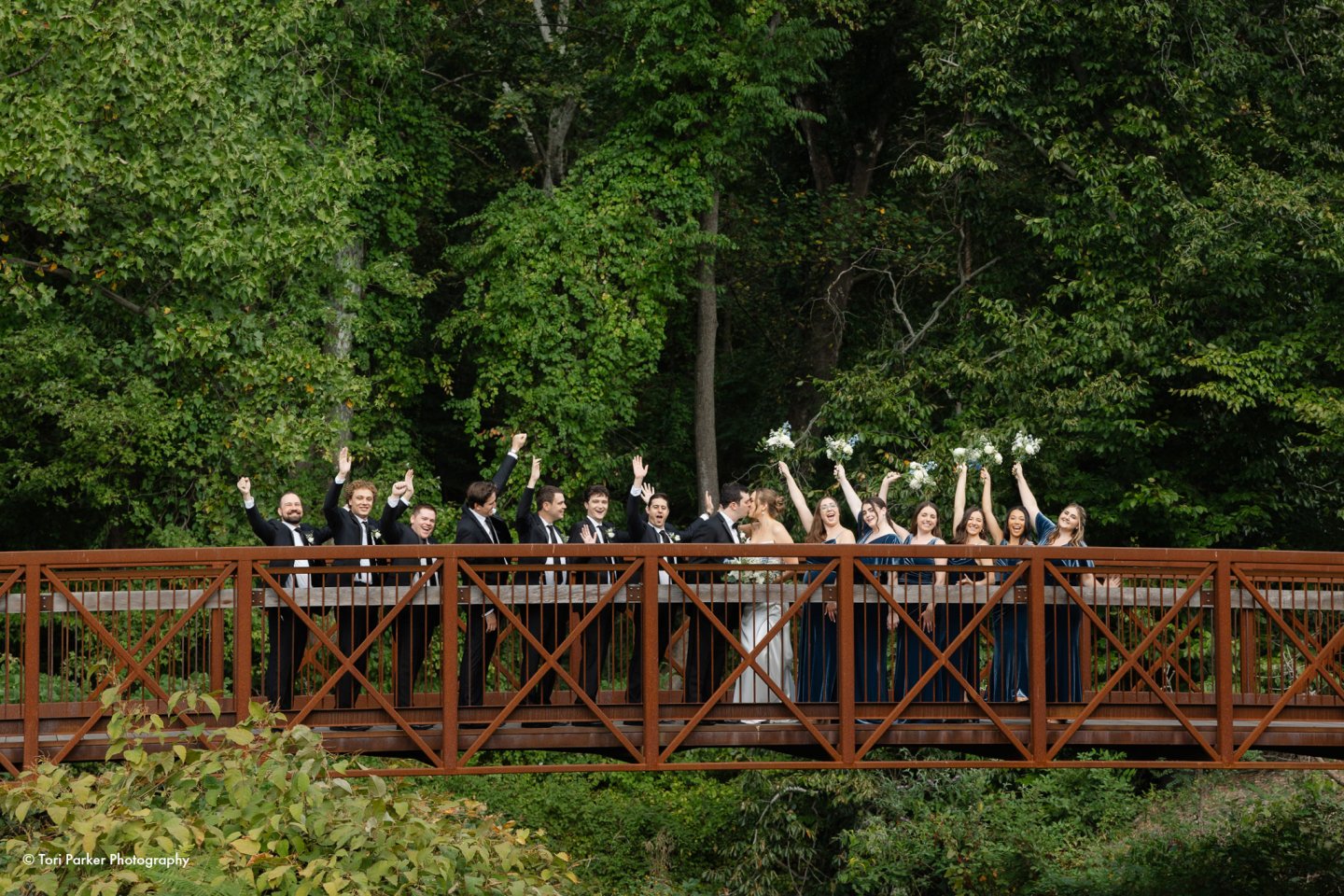 Wedding party celebrating on a wooden bridge amidst lush greenery.