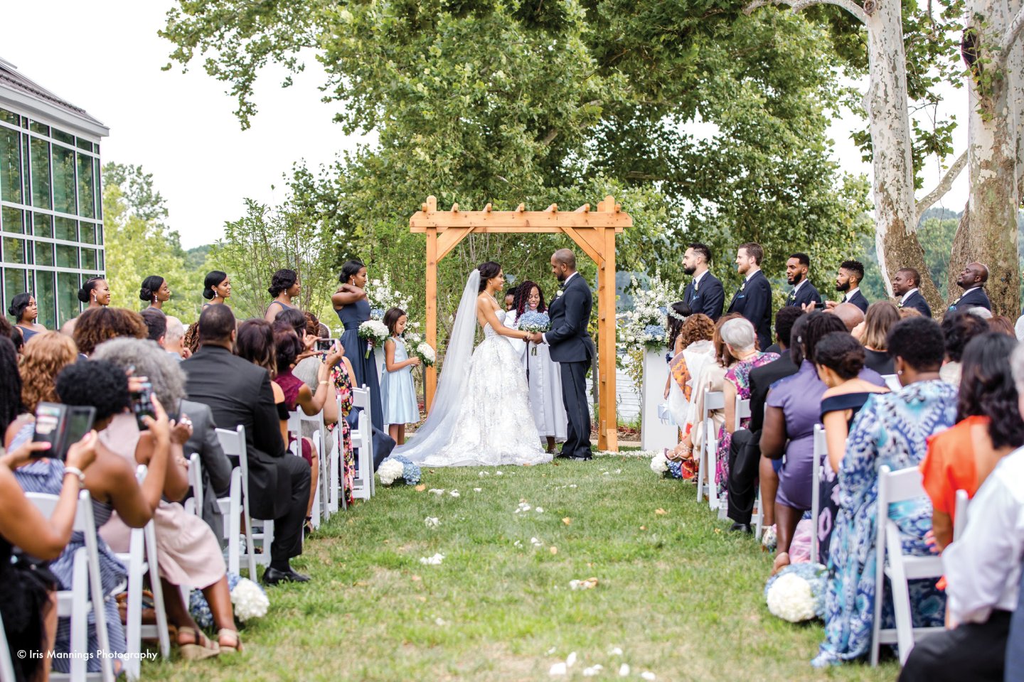 Wedding ceremony outdoors, couple under wooden arch, guests seated on either side.