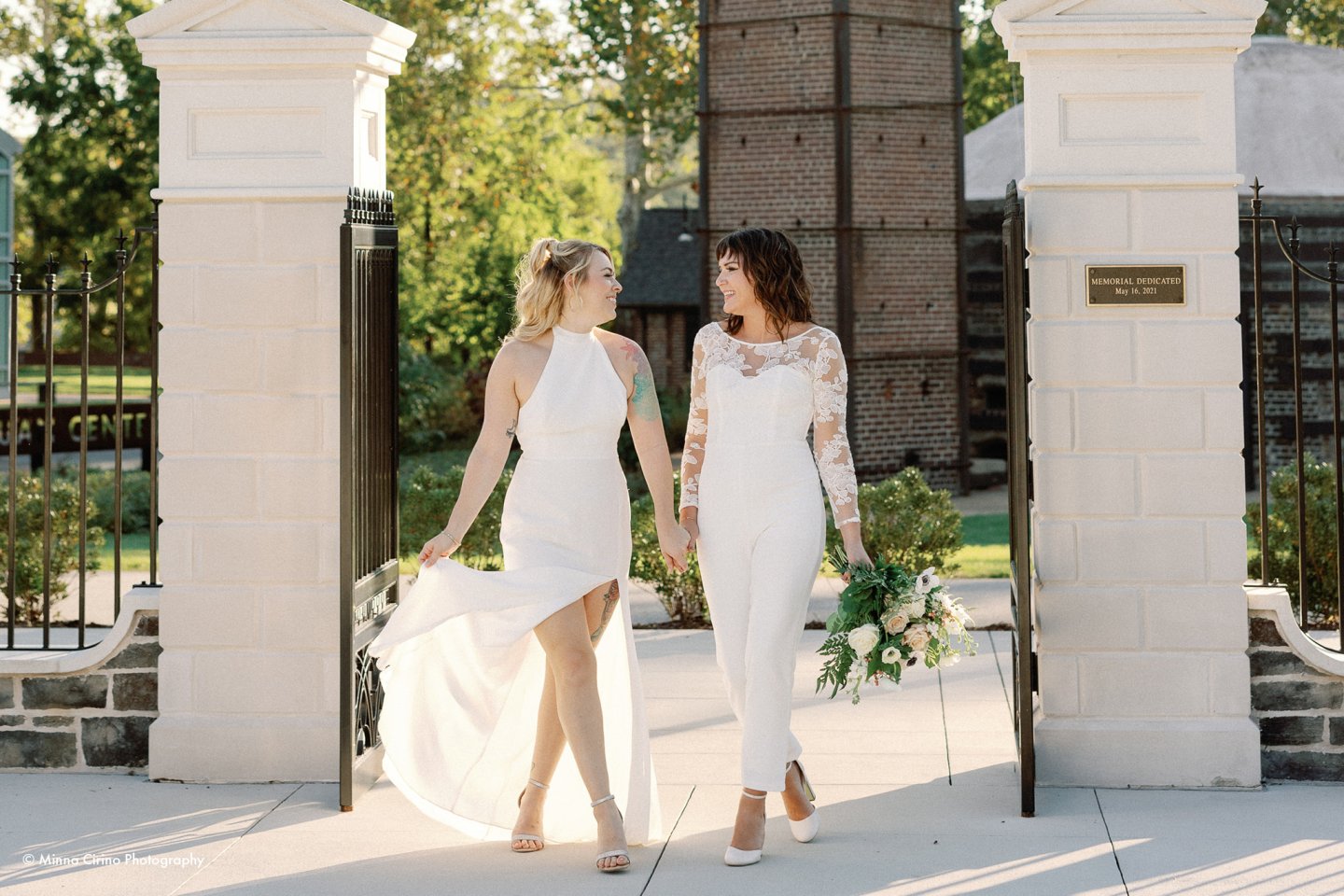 Two women in white wedding dresses walk hand in hand outdoors.