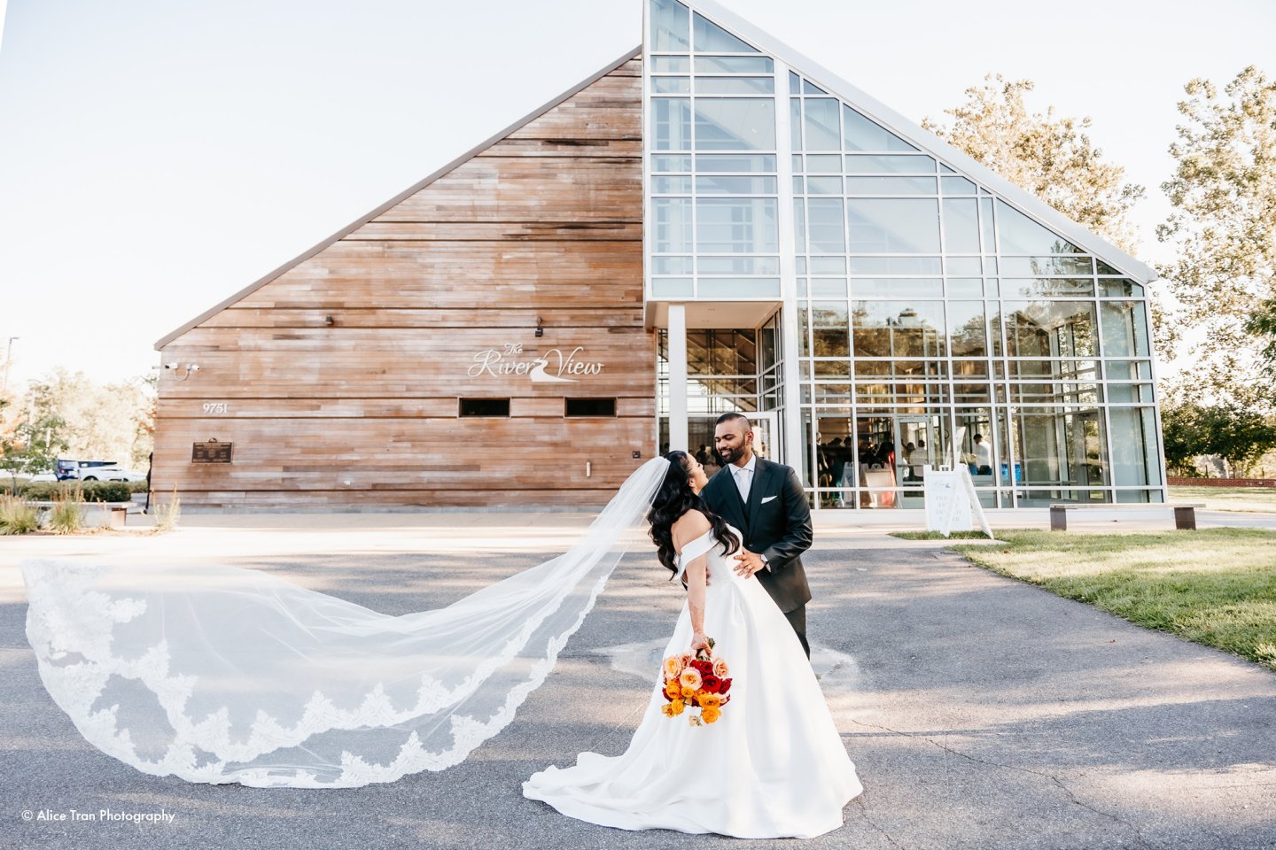 Bride and groom embrace, her veil flowing, in front of a modern building.