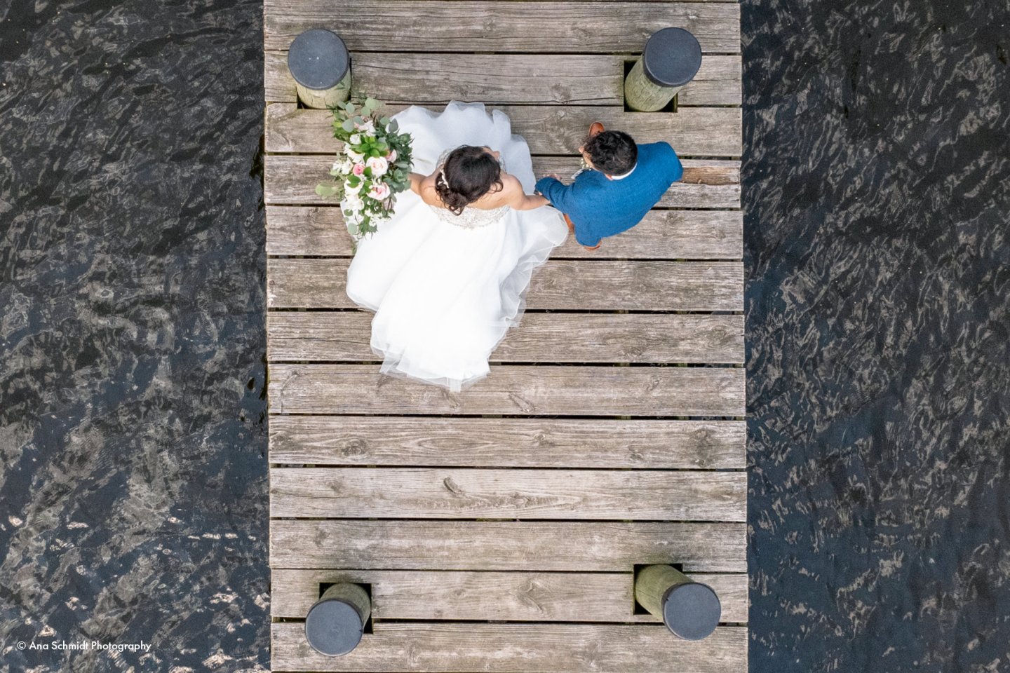 Bride and groom sitting on a wooden pier over water, seen from above.