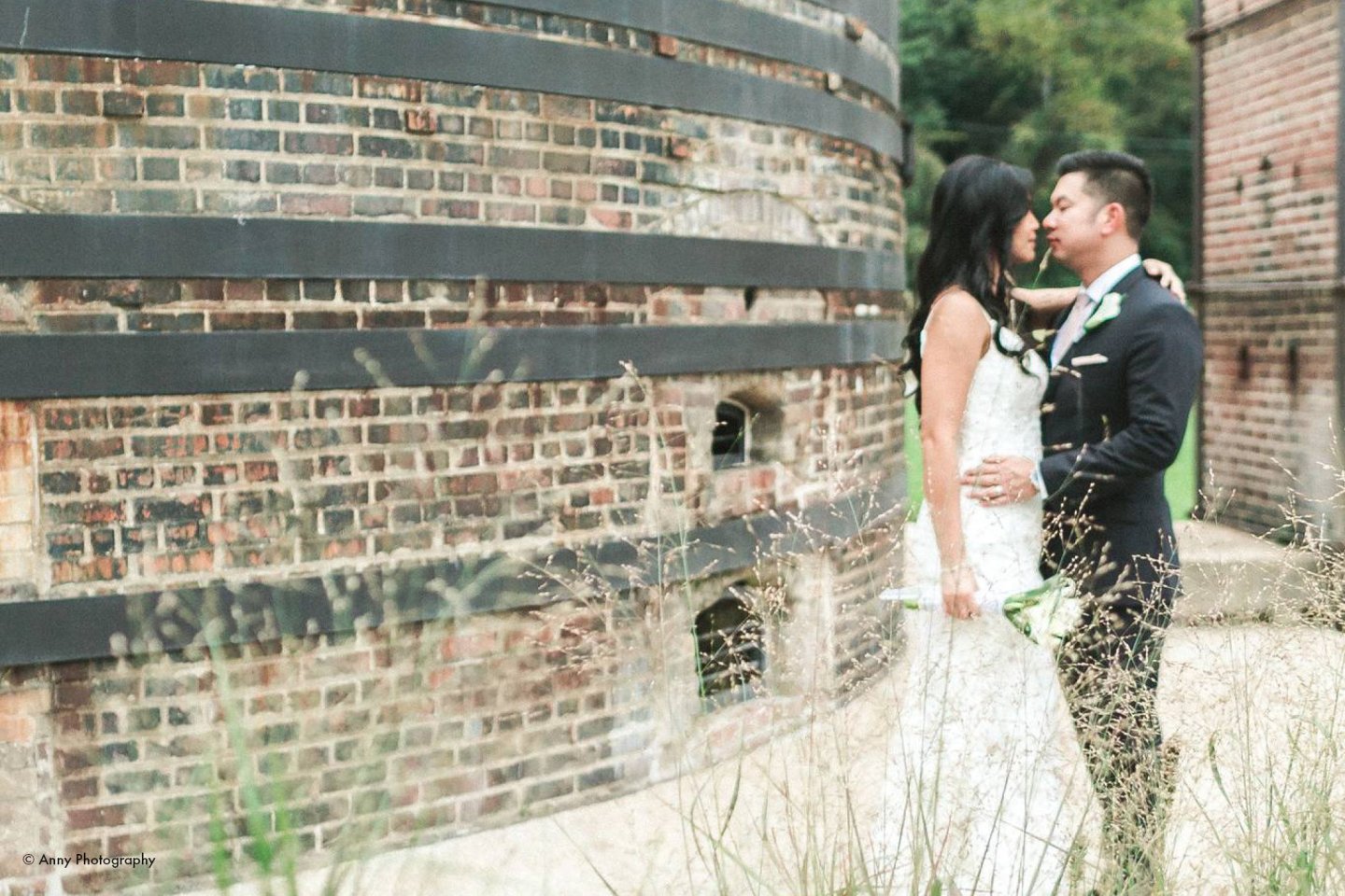 Bride and groom embracing near a brick structure outdoors.