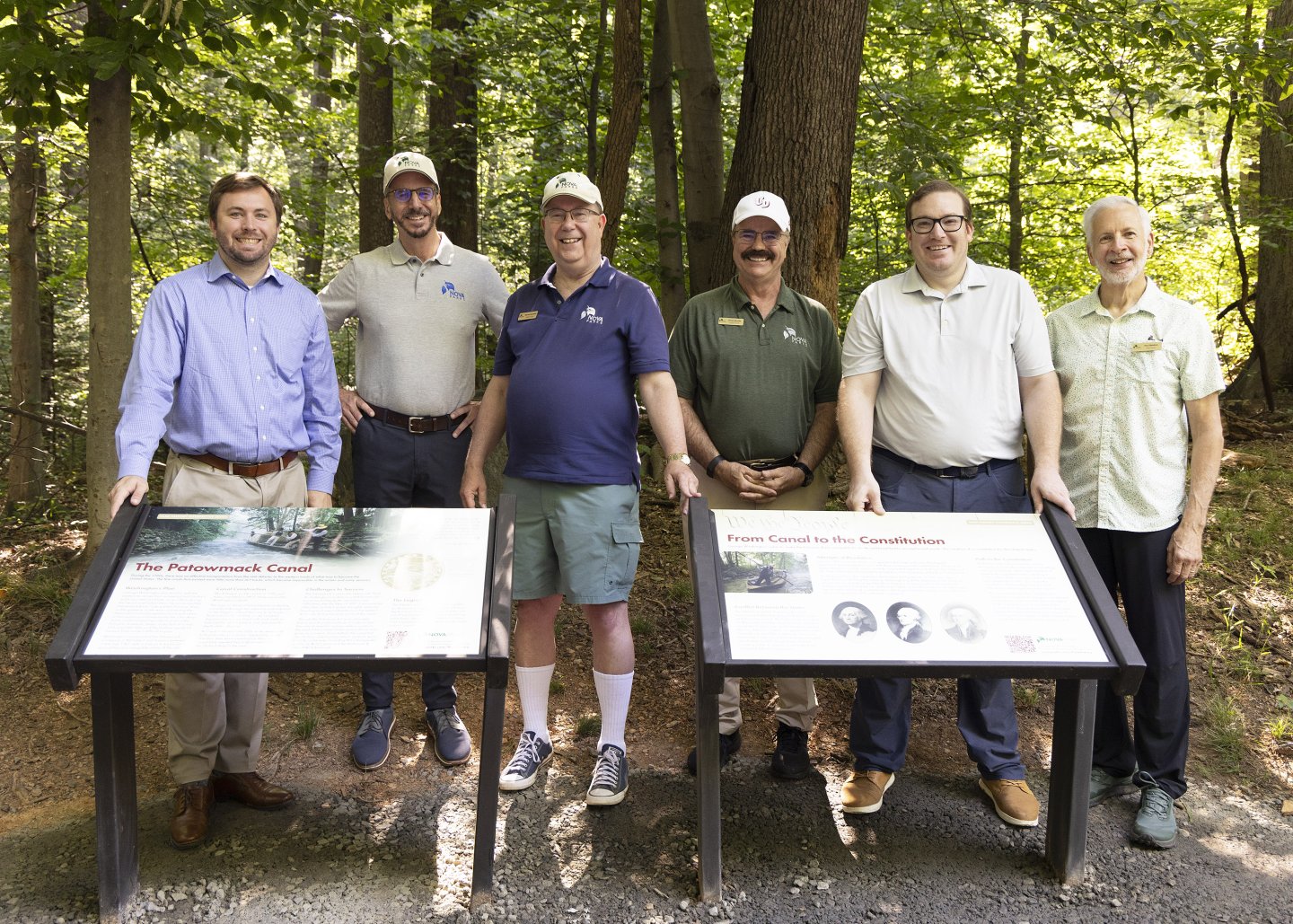 Six men standing outdoors beside informational signs in a forest setting.
