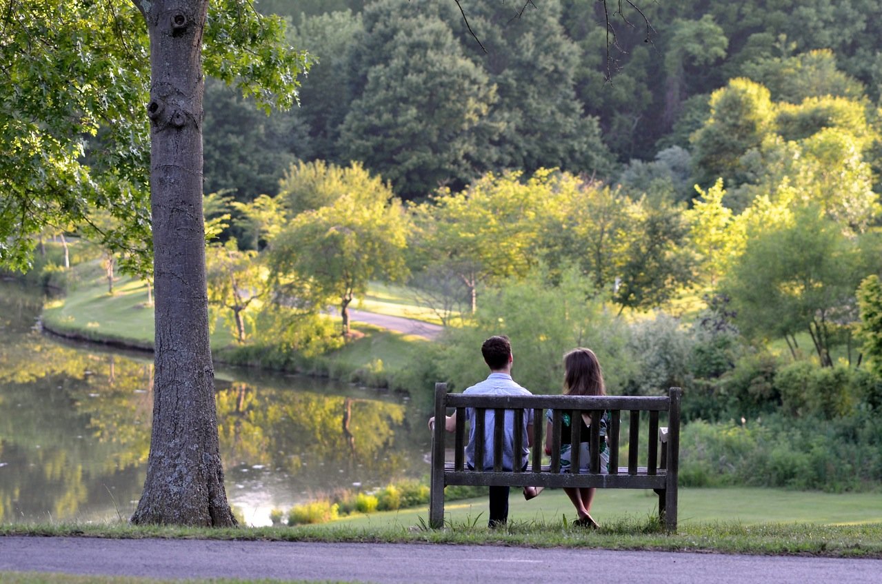 A couple sits on a bench overlooking a serene, tree-lined lake.