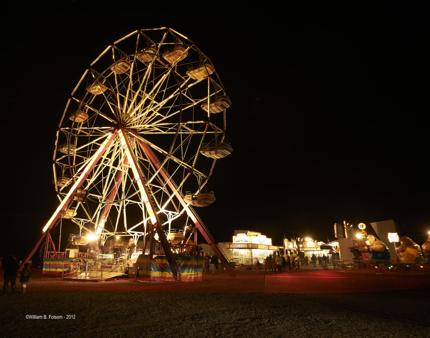 Ferris wheel at night, brightly lit, with nearby carnival stands.