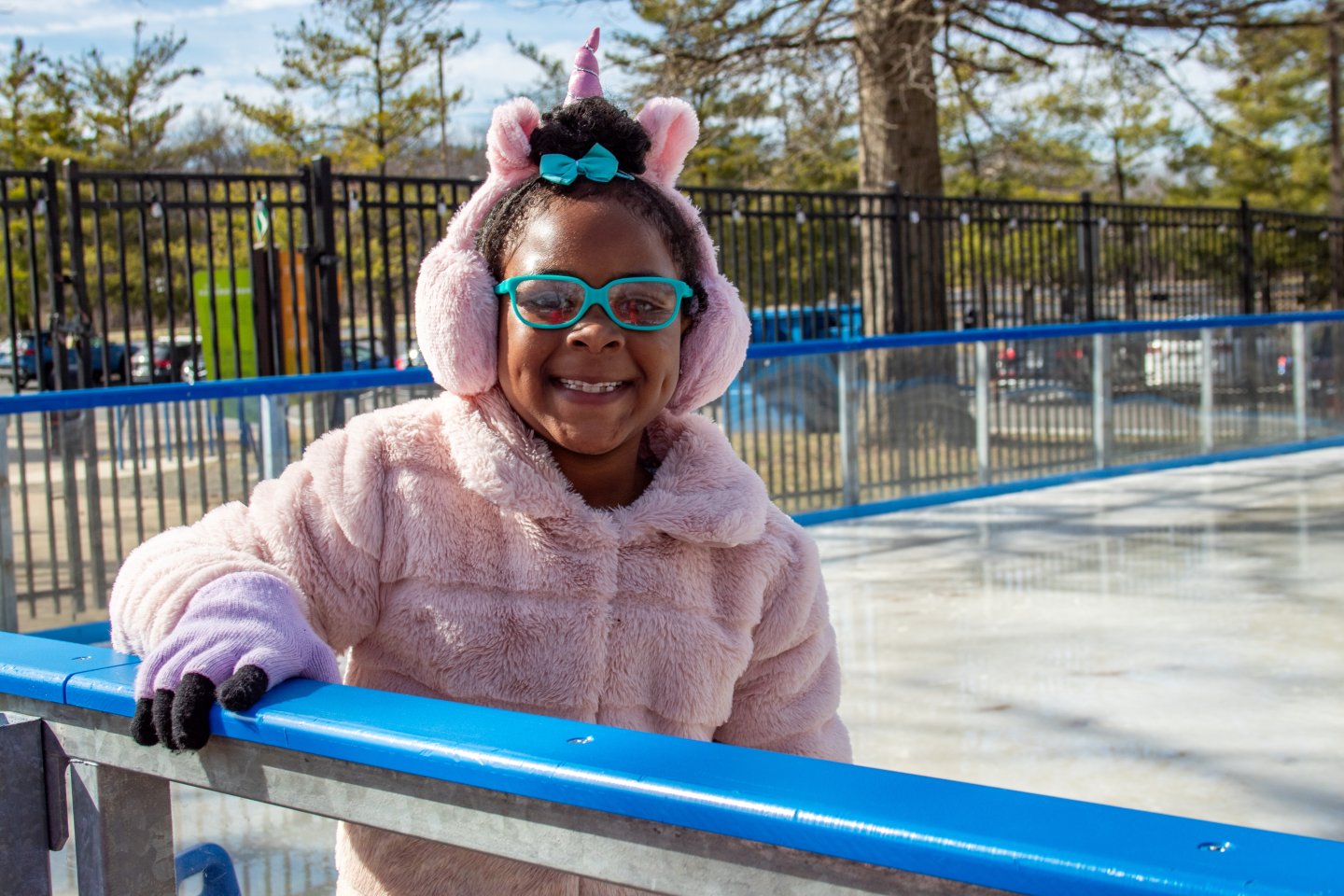 Smiling girl in winter coat and unicorn earmuffs at an outdoor ice rink.