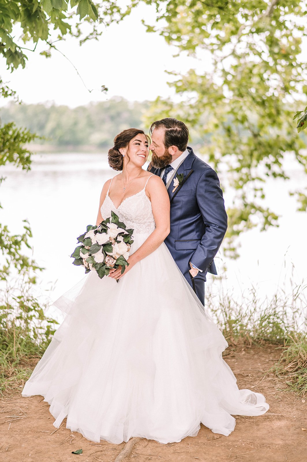 Bride and groom smiling by a lake, surrounded by greenery.