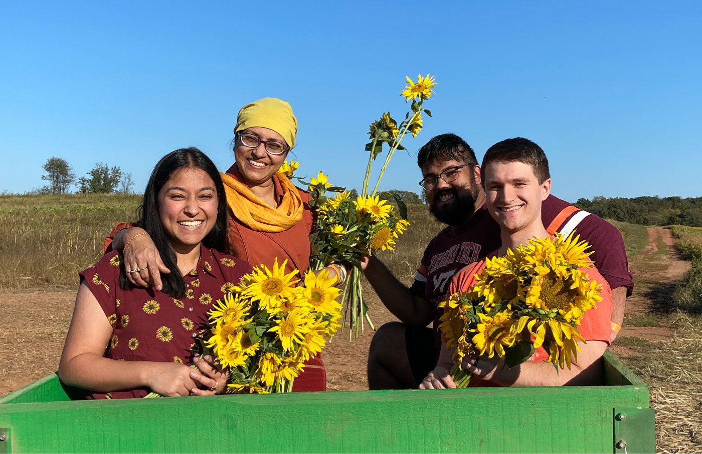 Four people holding sunflowers in a field, smiling, under a clear blue sky.