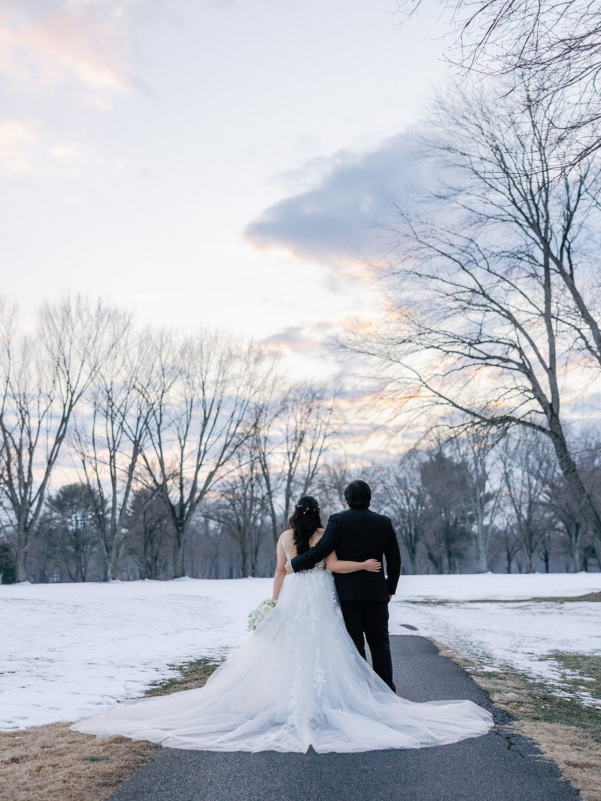 Couple in wedding attire walking on a snowy path, trees in background.