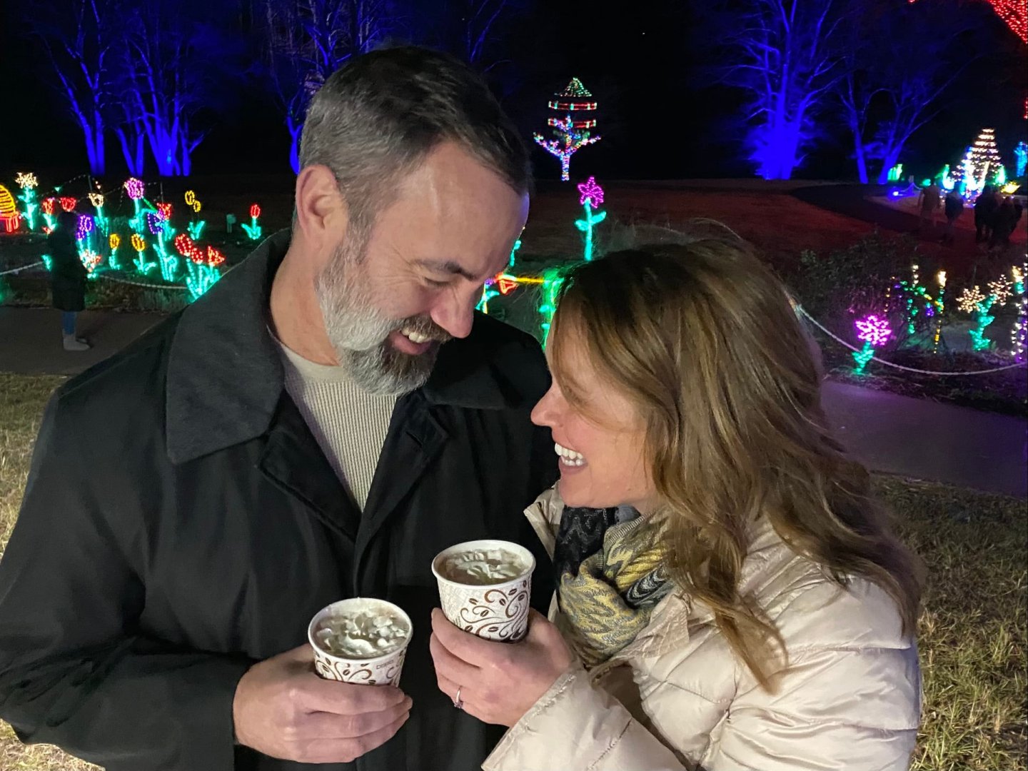 Couple smiling with hot drinks, colorful lights in background.