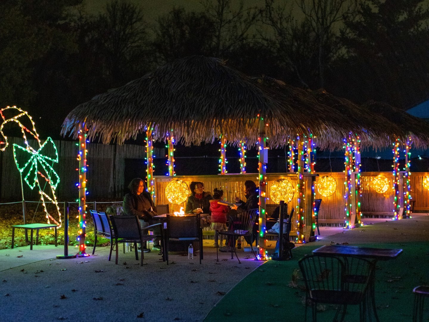Tiki hut decorated with colorful lights, people sitting around a fire pit at night.
