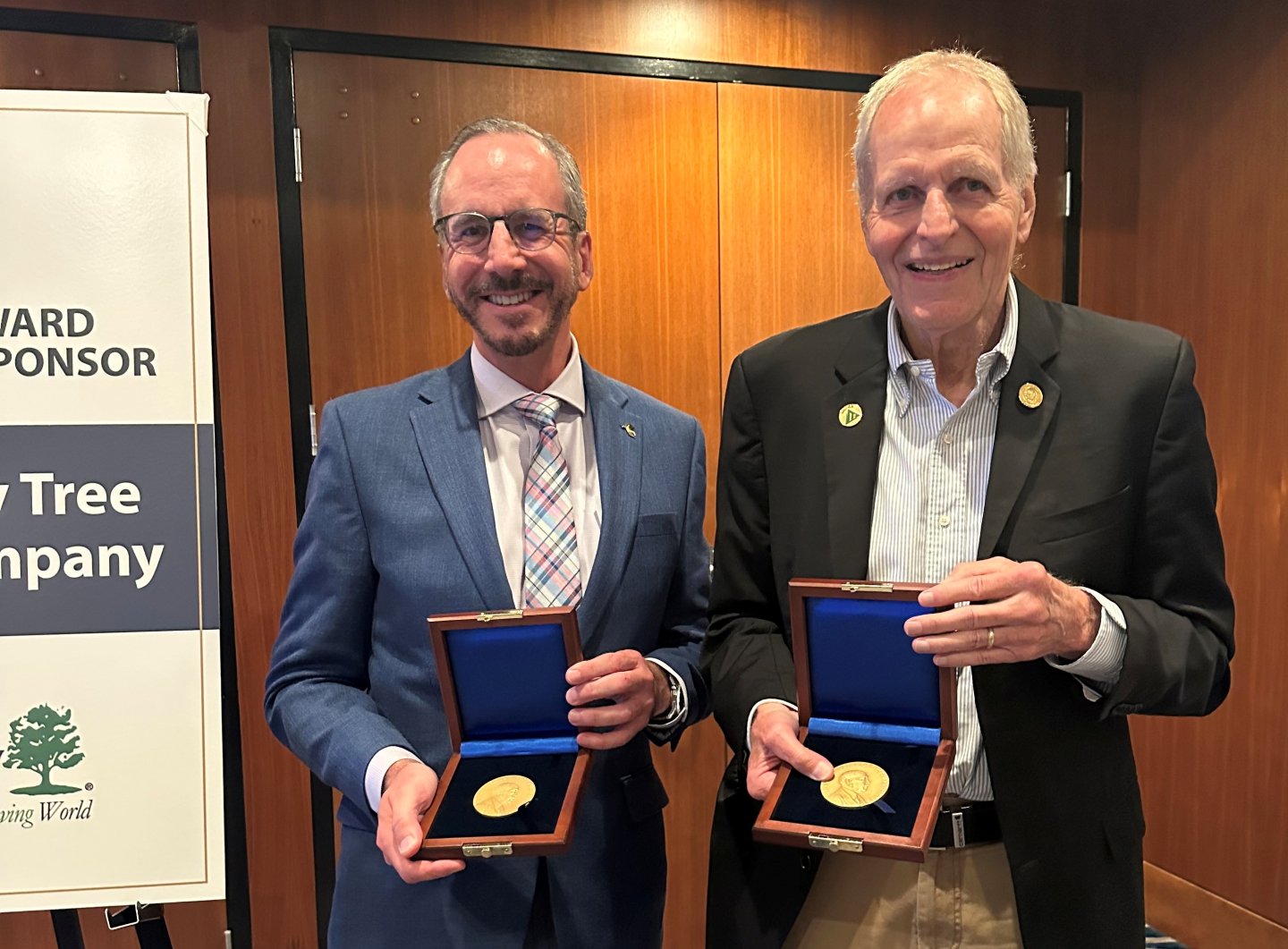 Two men in suits smiling, holding awards in wooden boxes indoors.