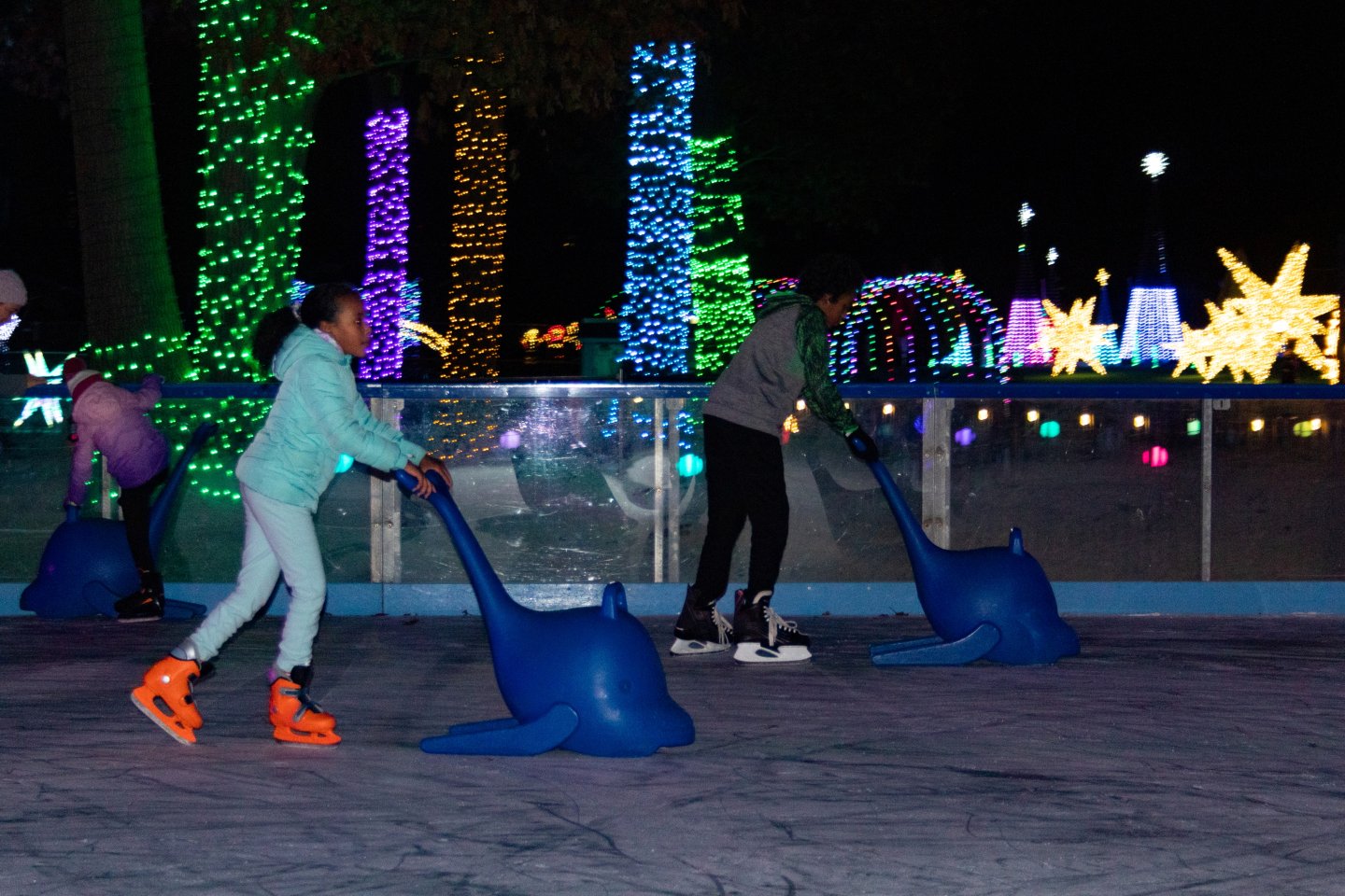 Kids ice skating at night with colorful lights and blue dolphin aids.