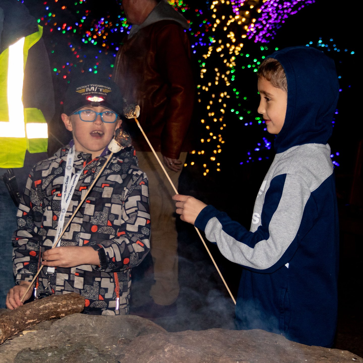 Two children roasting marshmallows at night near colorful lights.