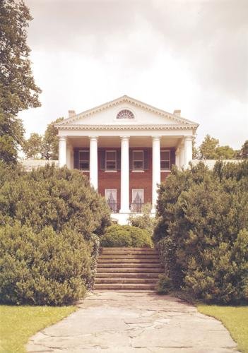 Historic mansion with white columns and red brick facade, surrounded by greenery.