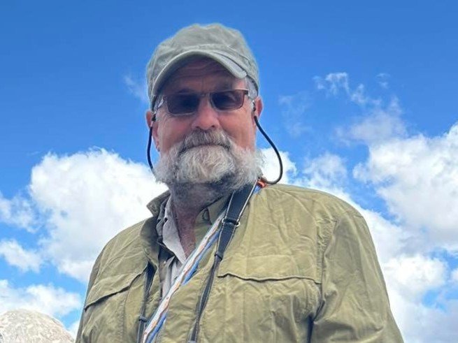 Man in outdoor clothing and cap against a blue sky with clouds.