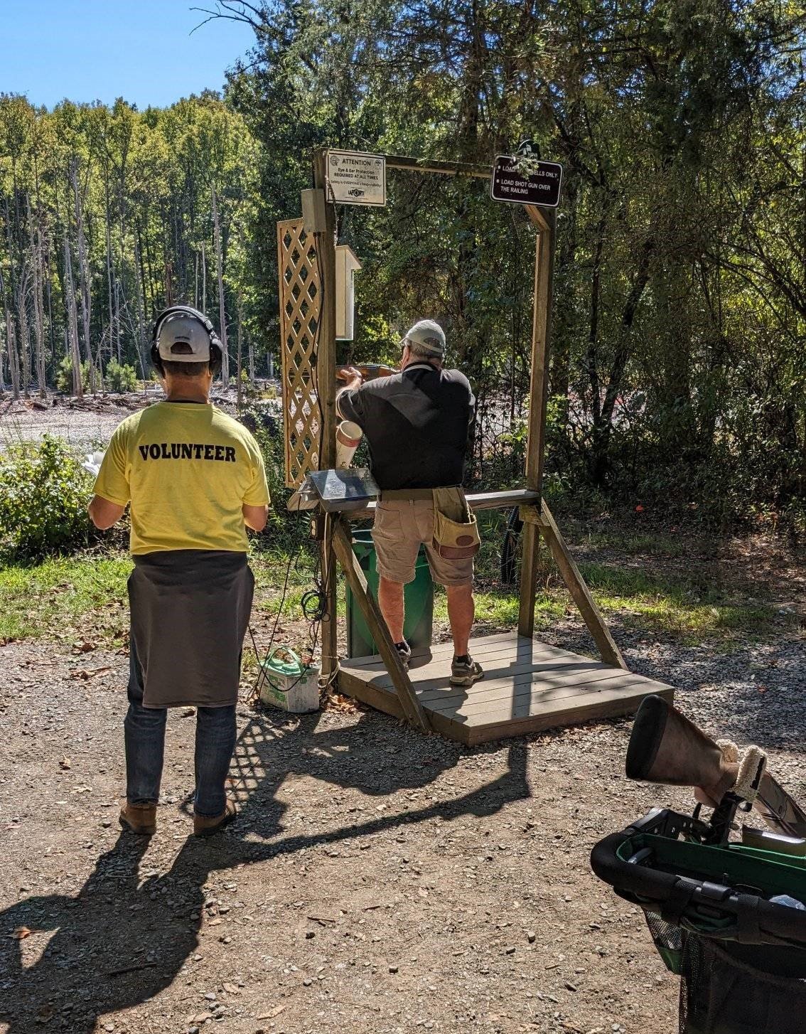 Two people at an outdoor shooting range, one aiming, one in a yellow volunteer shirt.