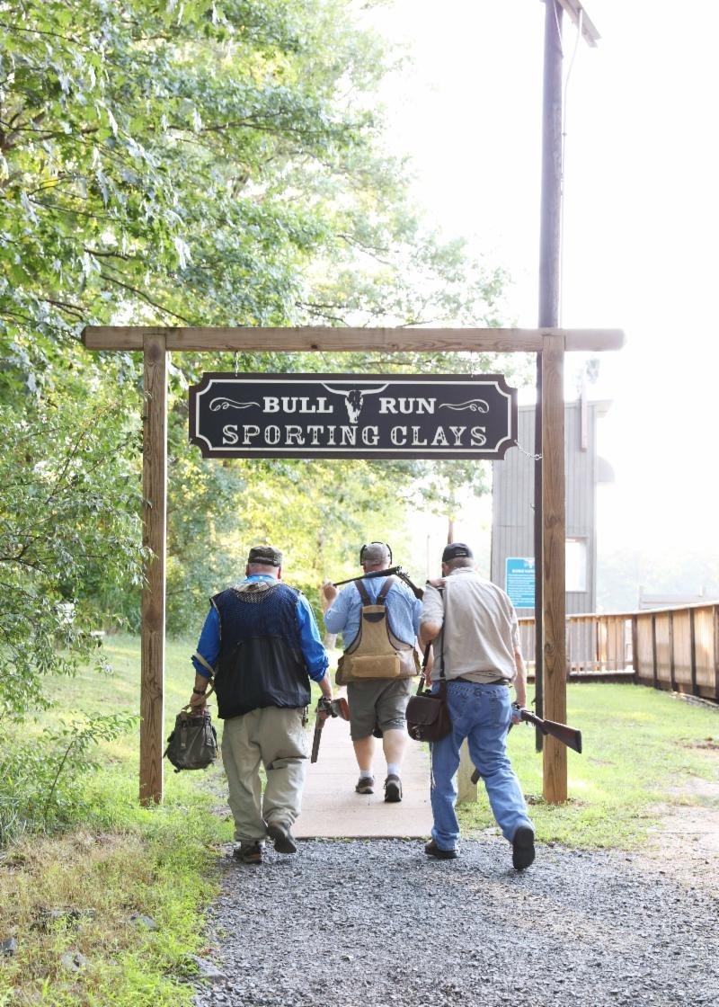Three people walking under a "Sporting Clays" entrance sign with shotguns and gear.