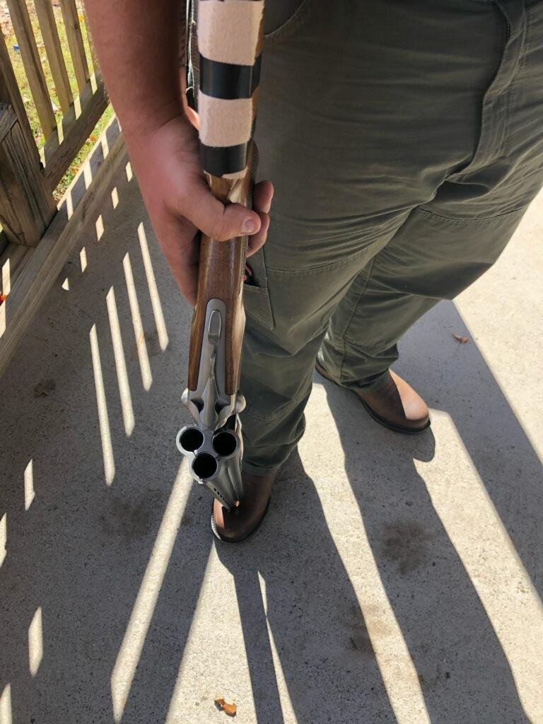 Person holding a shotgun, standing on a sunlit porch.
