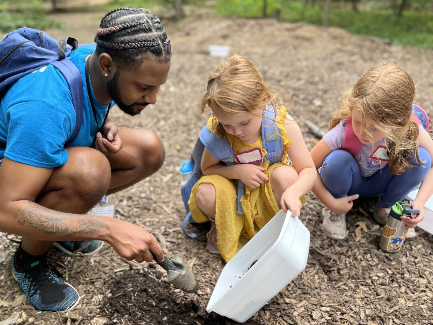Man and two children examining soil with a spade outdoors.
