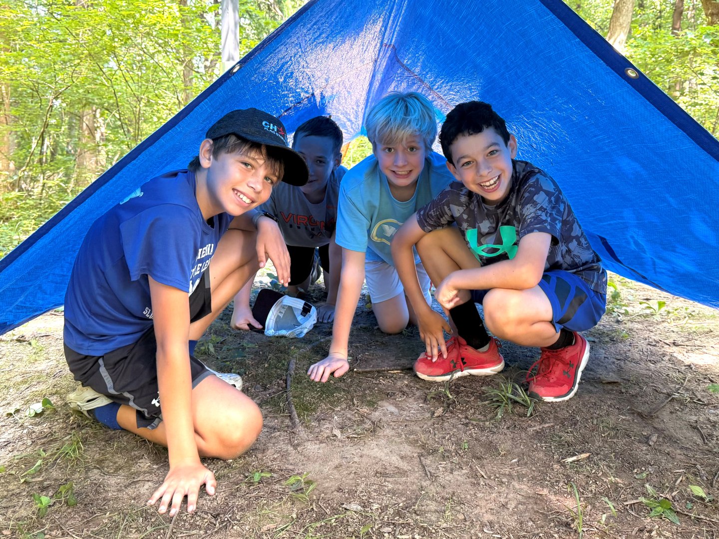 Boys smiling under a blue tarp tent in a forest setting.