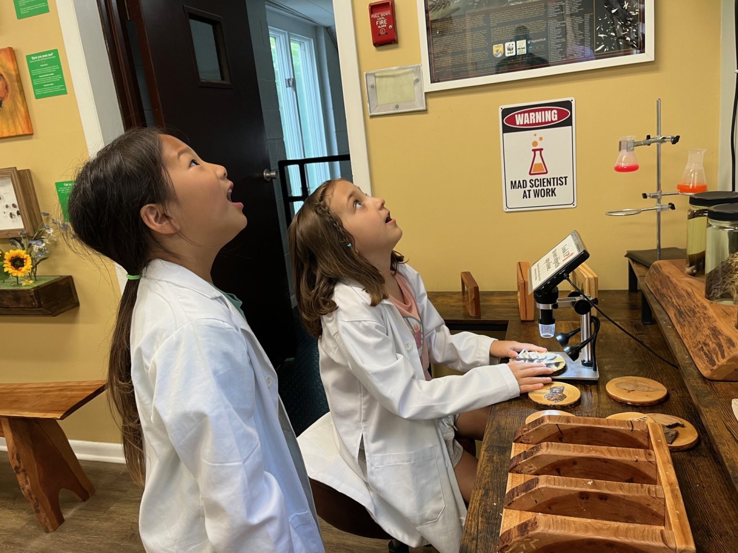 Two kids in lab coats looking up, surprised in a science room.