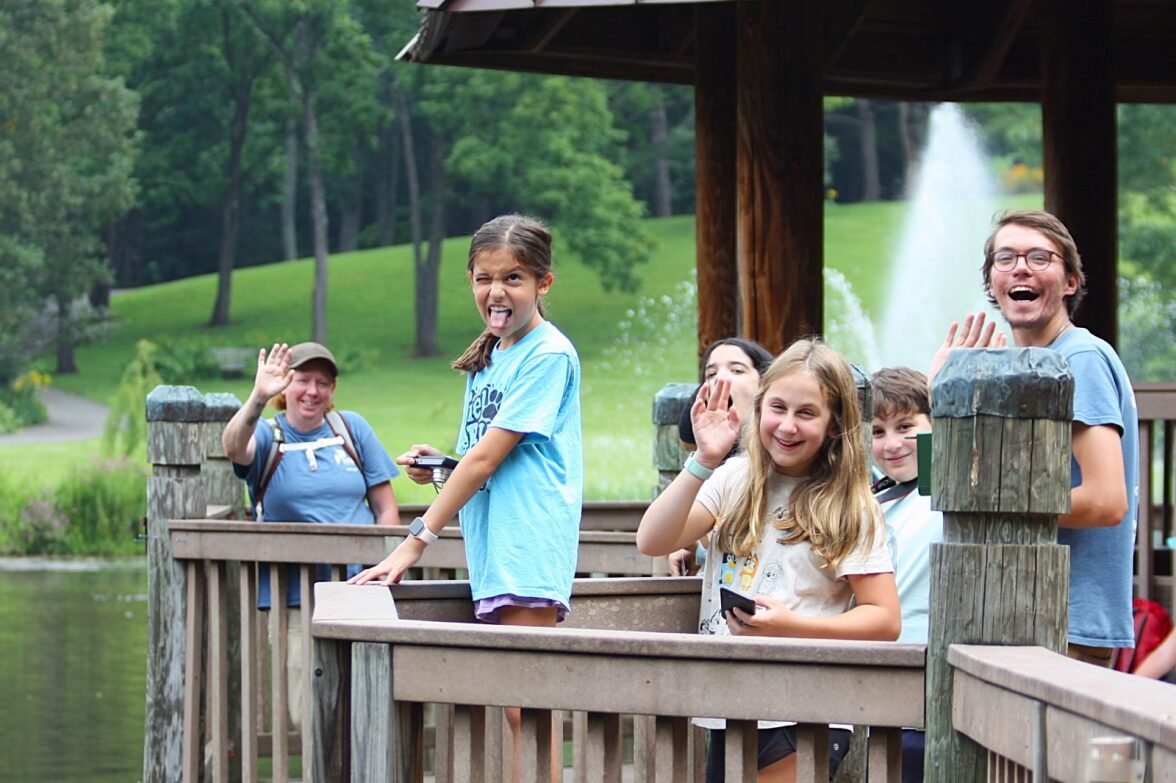 Group of people smiling and waving on a wooden deck by a pond with fountain.