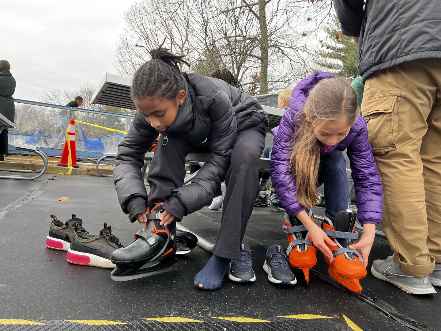 Two children sitting and putting on ice skates outdoors.