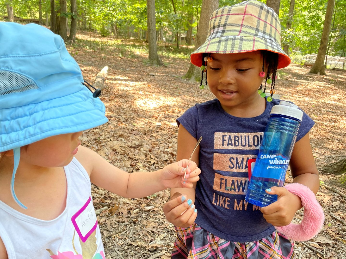 Two children outdoors, wearing hats, one holding a bottle, surrounded by trees.