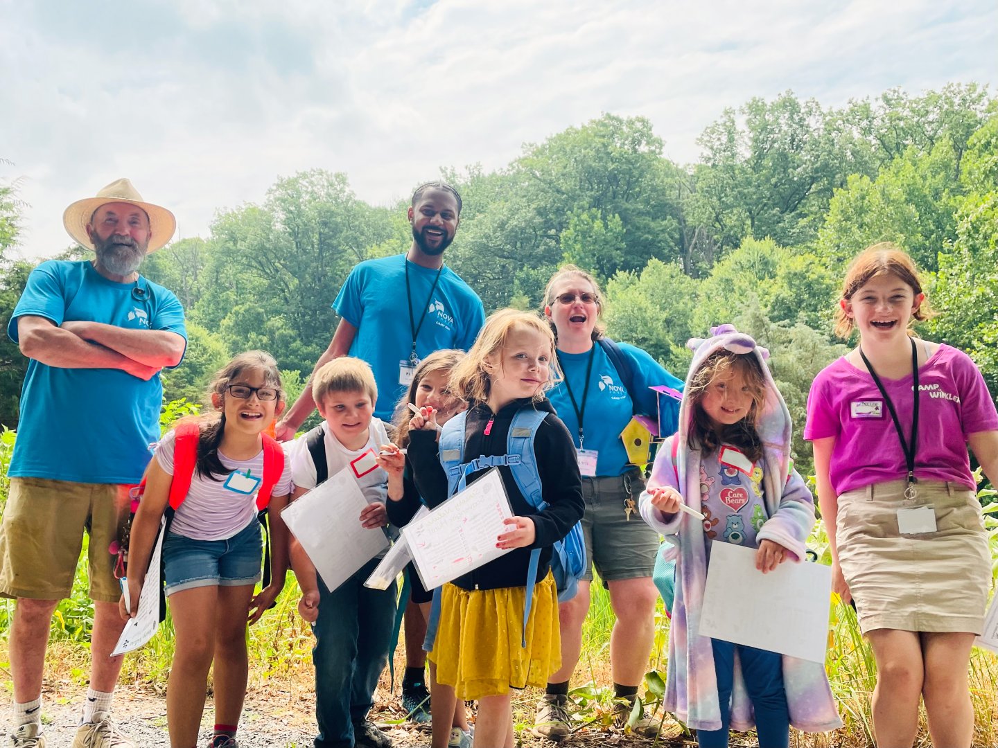 Kids and adults outdoors holding papers, smiling in a green park setting.