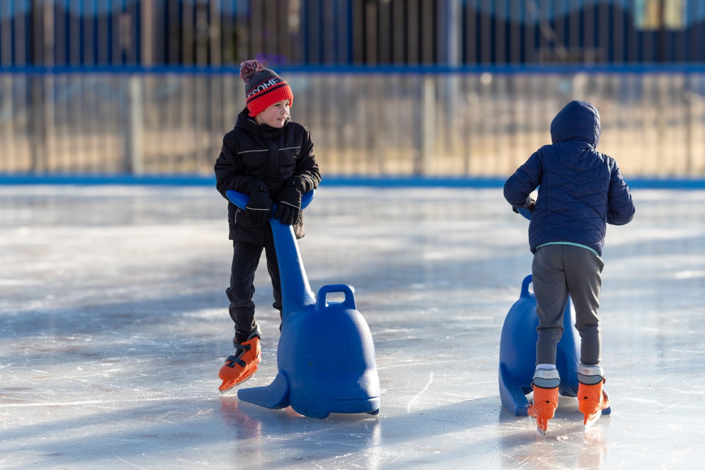 Two children ice skating with blue supports on a rink.