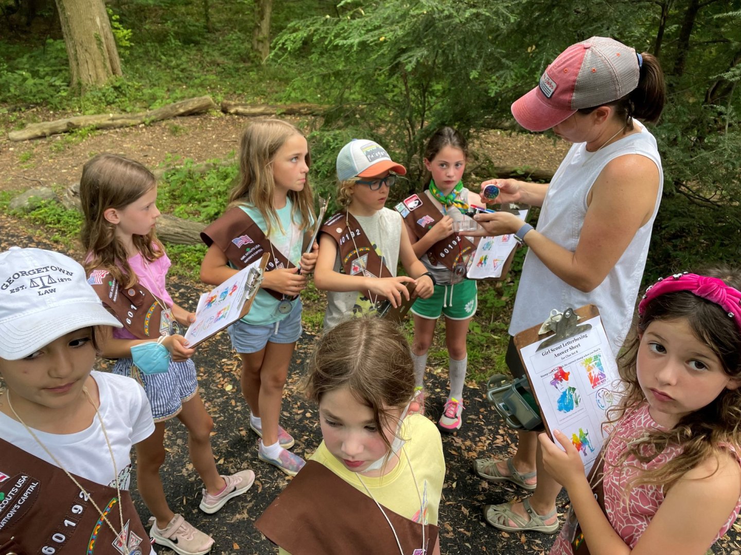 Girls in uniforms listen to a leader in a forest, holding activity sheets.