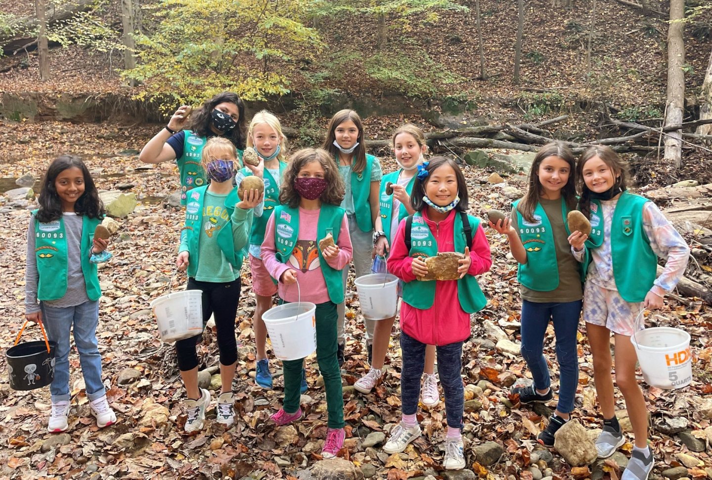 A group of smiling girls with buckets in a forest setting, wearing green vests.
