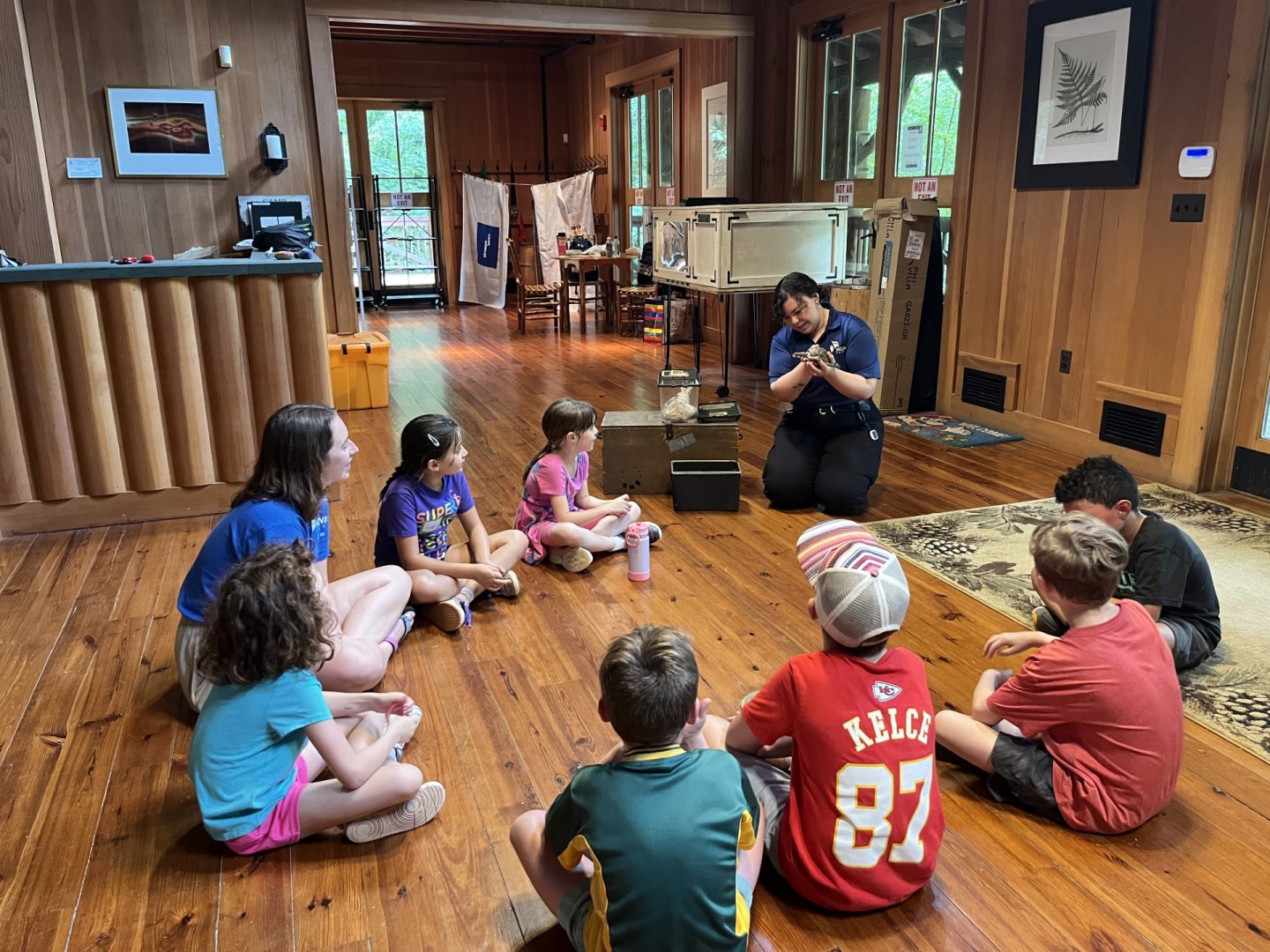 Kids sitting in a circle on a wooden floor, listening to an instructor indoors.