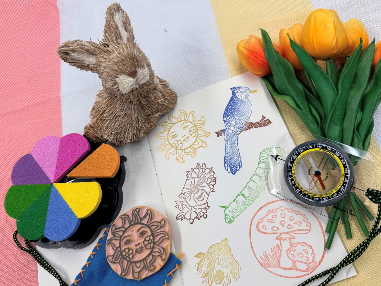 Craft items include a rabbit, ink pad, stamps, tulips, and a compass on a colorful background.