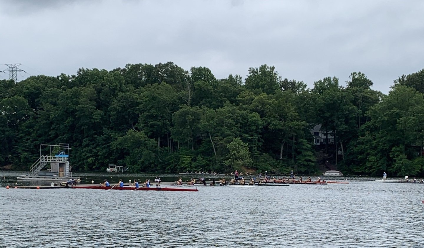 Rowers on a river with a forested background under a cloudy sky.