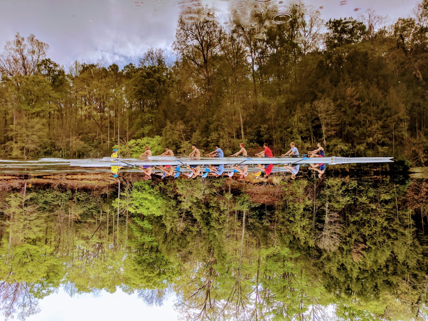 Rowing team paddles on calm river, surrounded by trees, reflections visible.