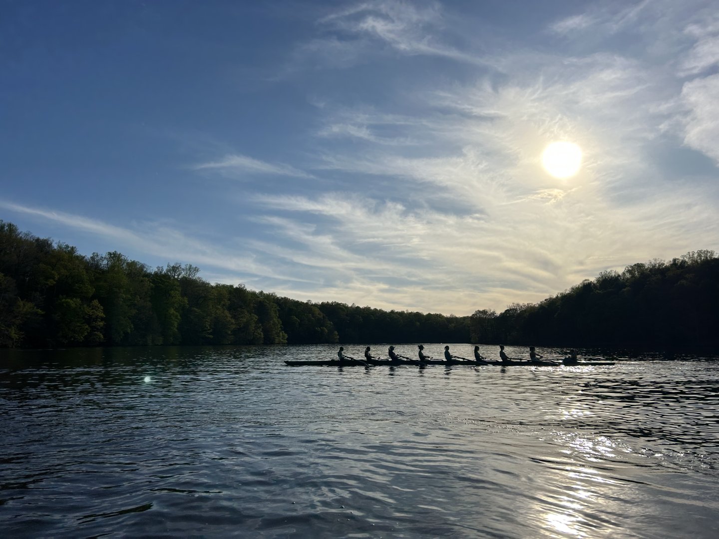 Rowers on a river at sunset, surrounded by trees and a bright sky.