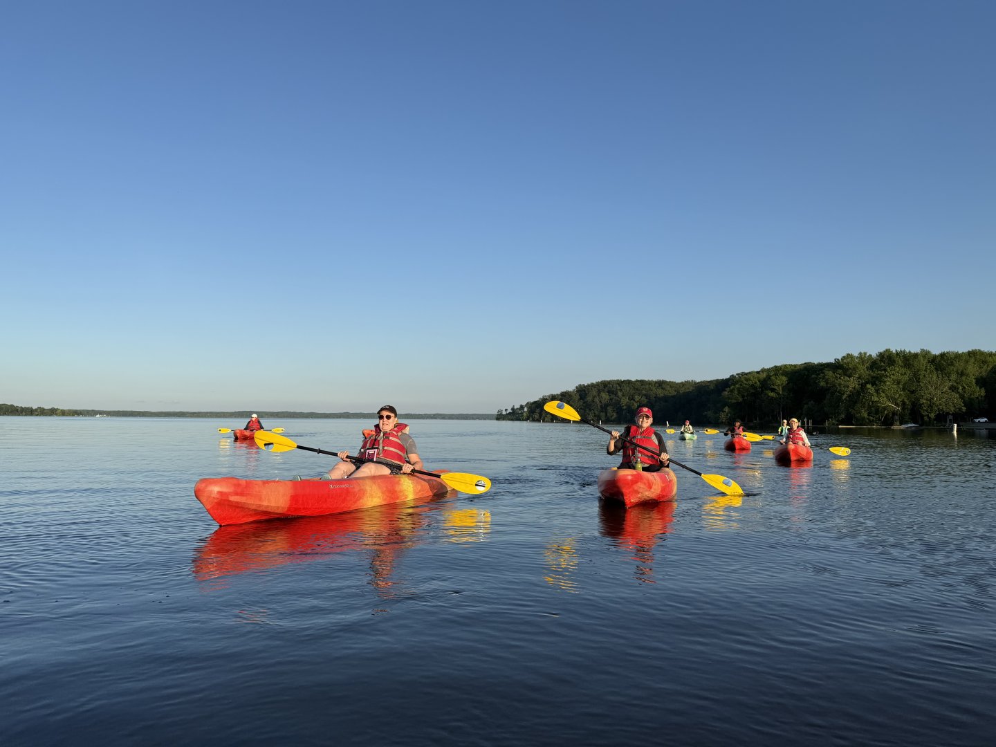 Kayakers in red kayaks paddle on a calm lake under a clear blue sky.