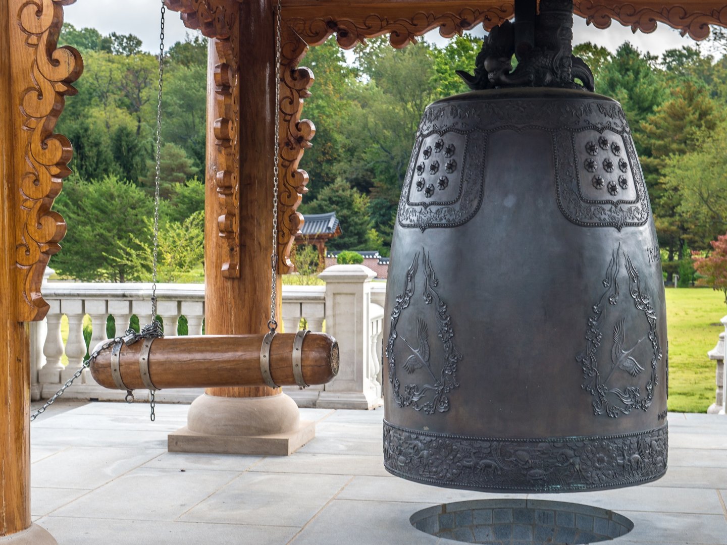 Large bronze bell under ornate wooden pavilion near lush garden.