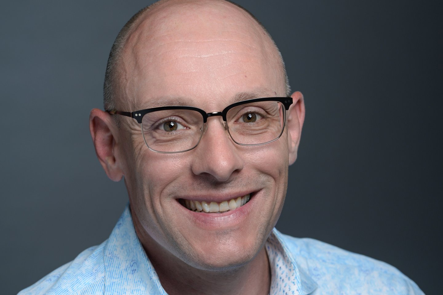 Man wearing glasses and a light blue shirt, smiling against a dark background.