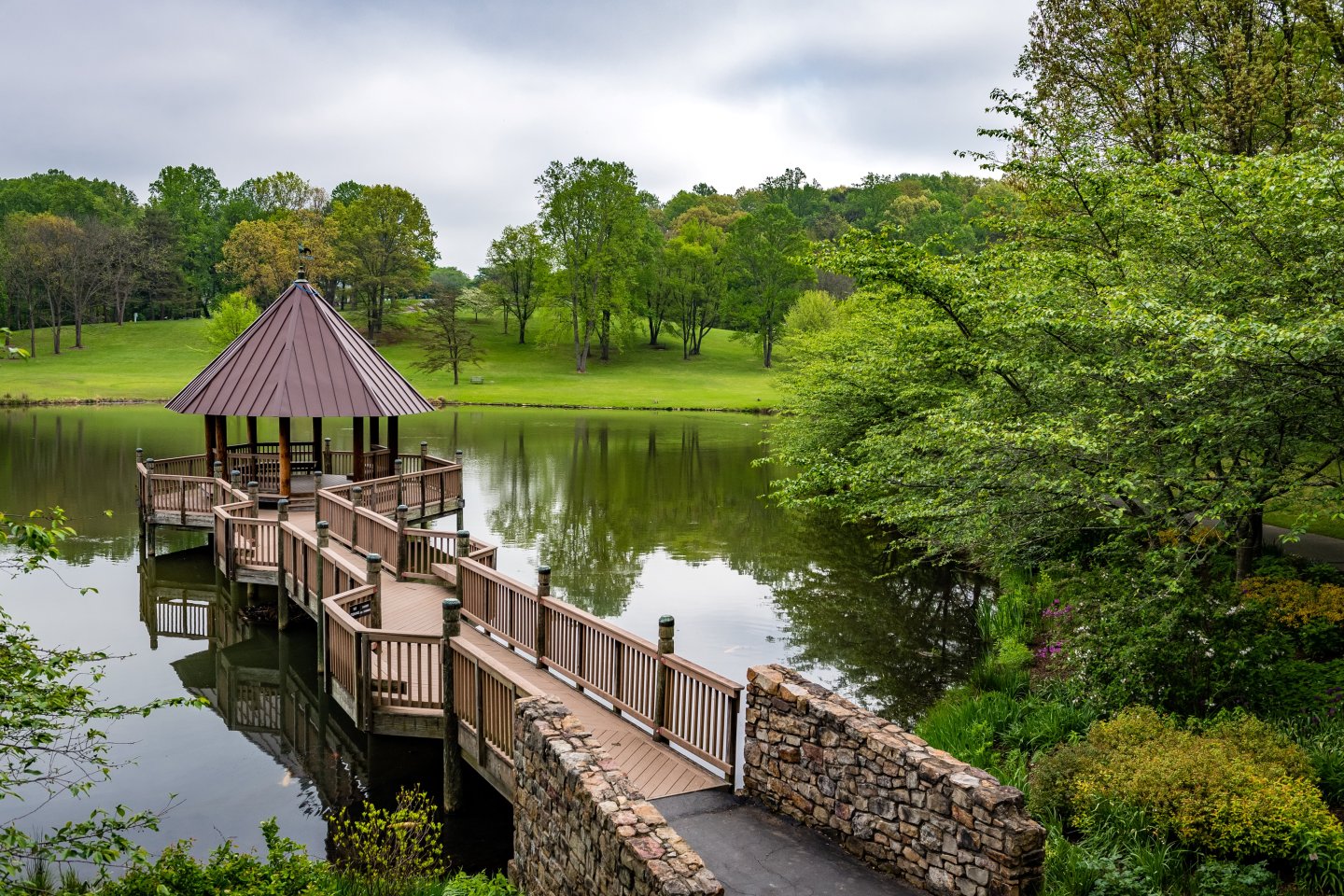 Pagoda on a lake with a wooden walkway, surrounded by lush green trees.
