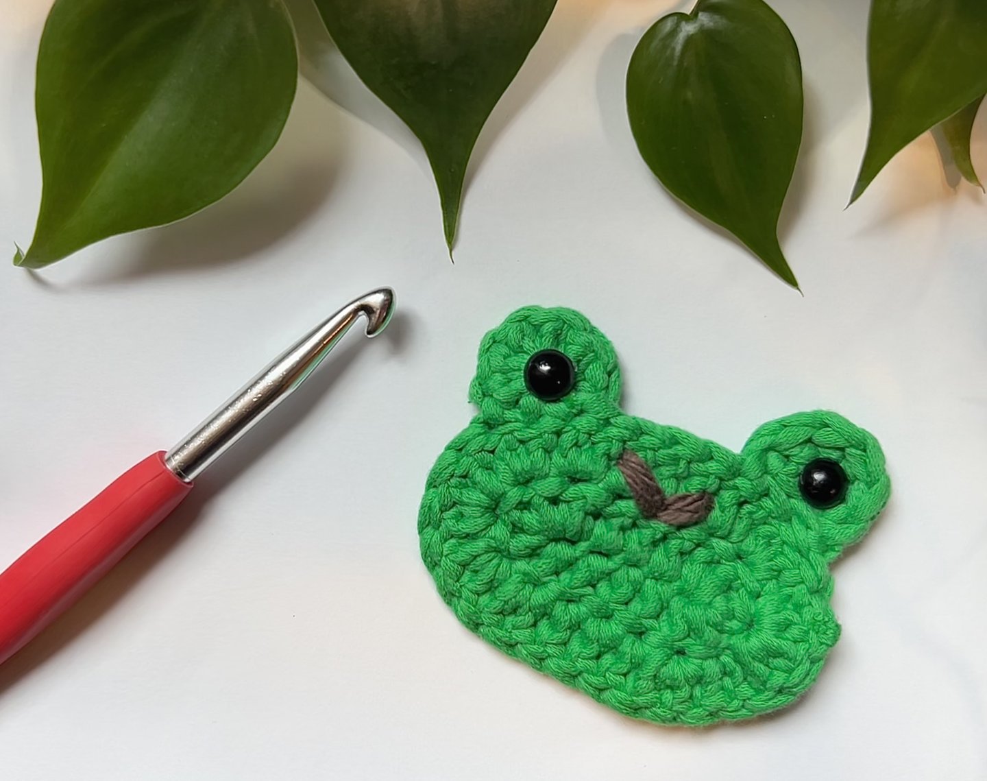 Crocheted green frog face with leaves and a crochet hook.