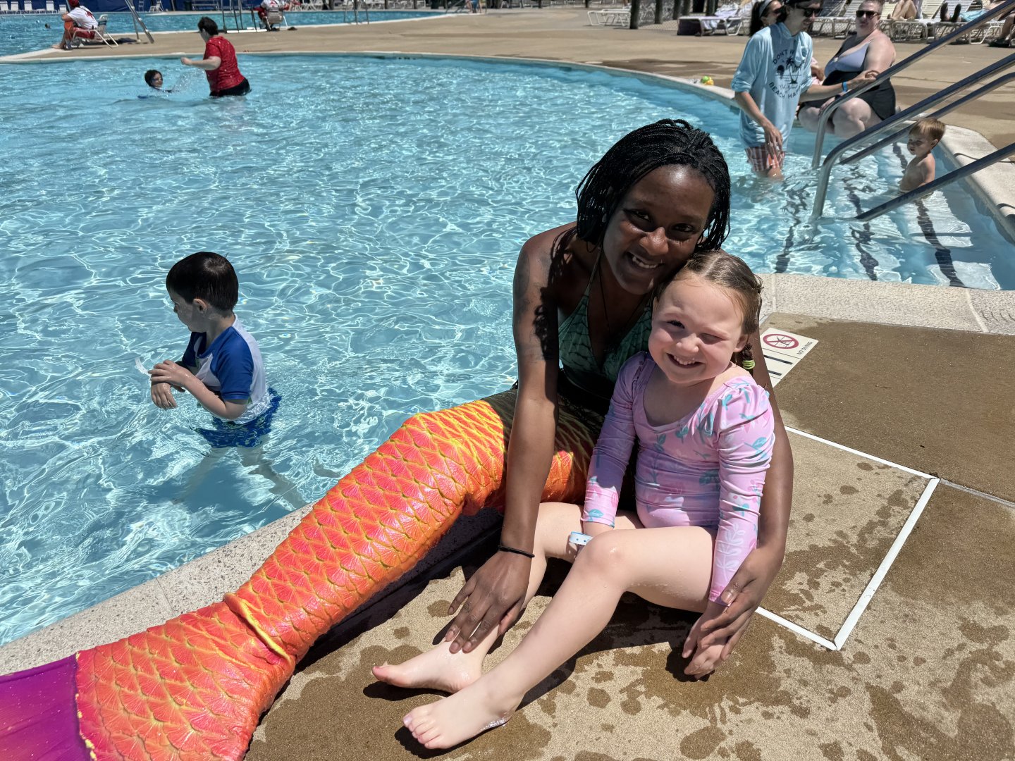 Woman with mermaid tail and child smiling by a pool.