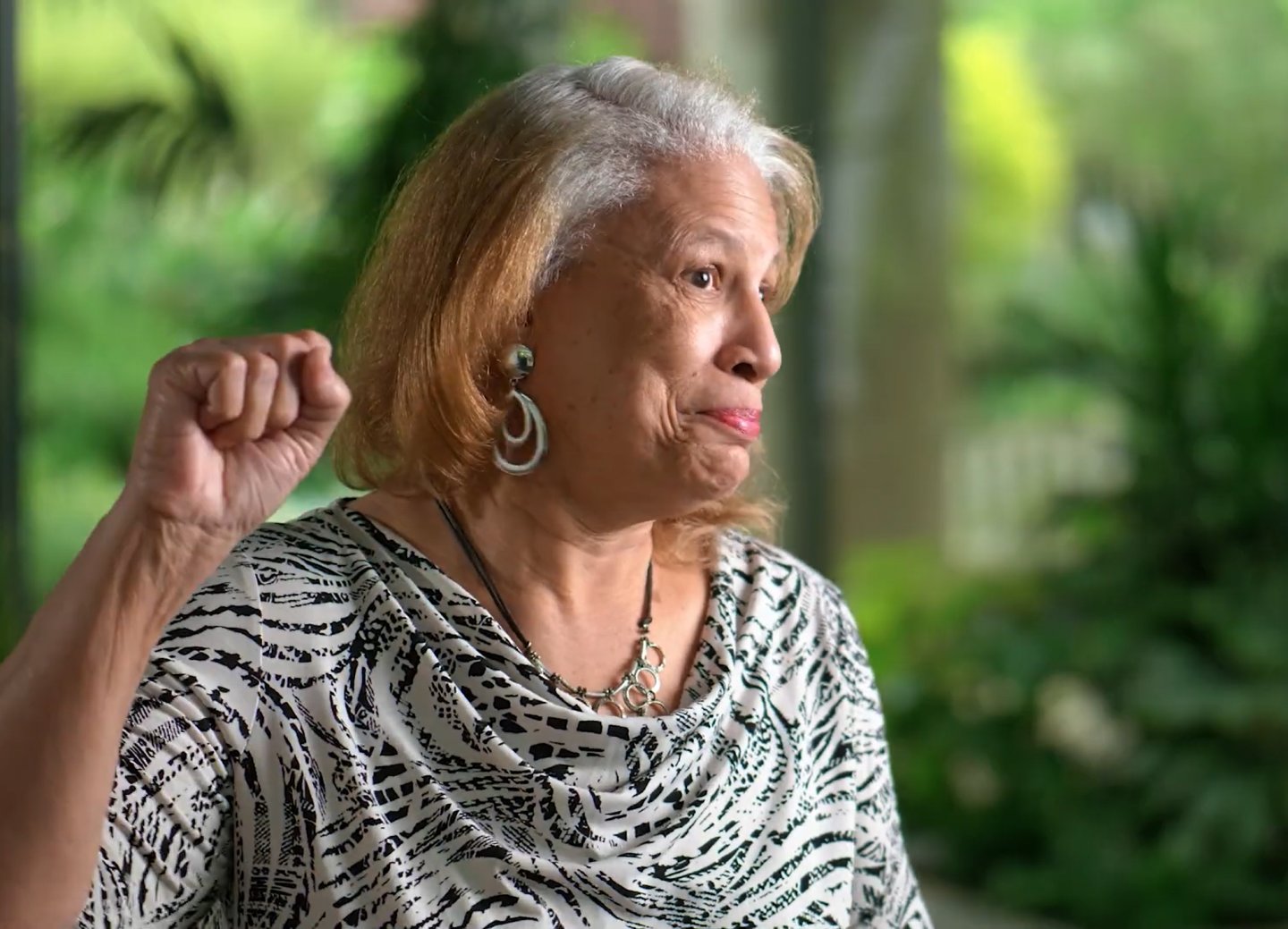  Woman gesturing, wearing a patterned blouse, in a lush garden setting.
