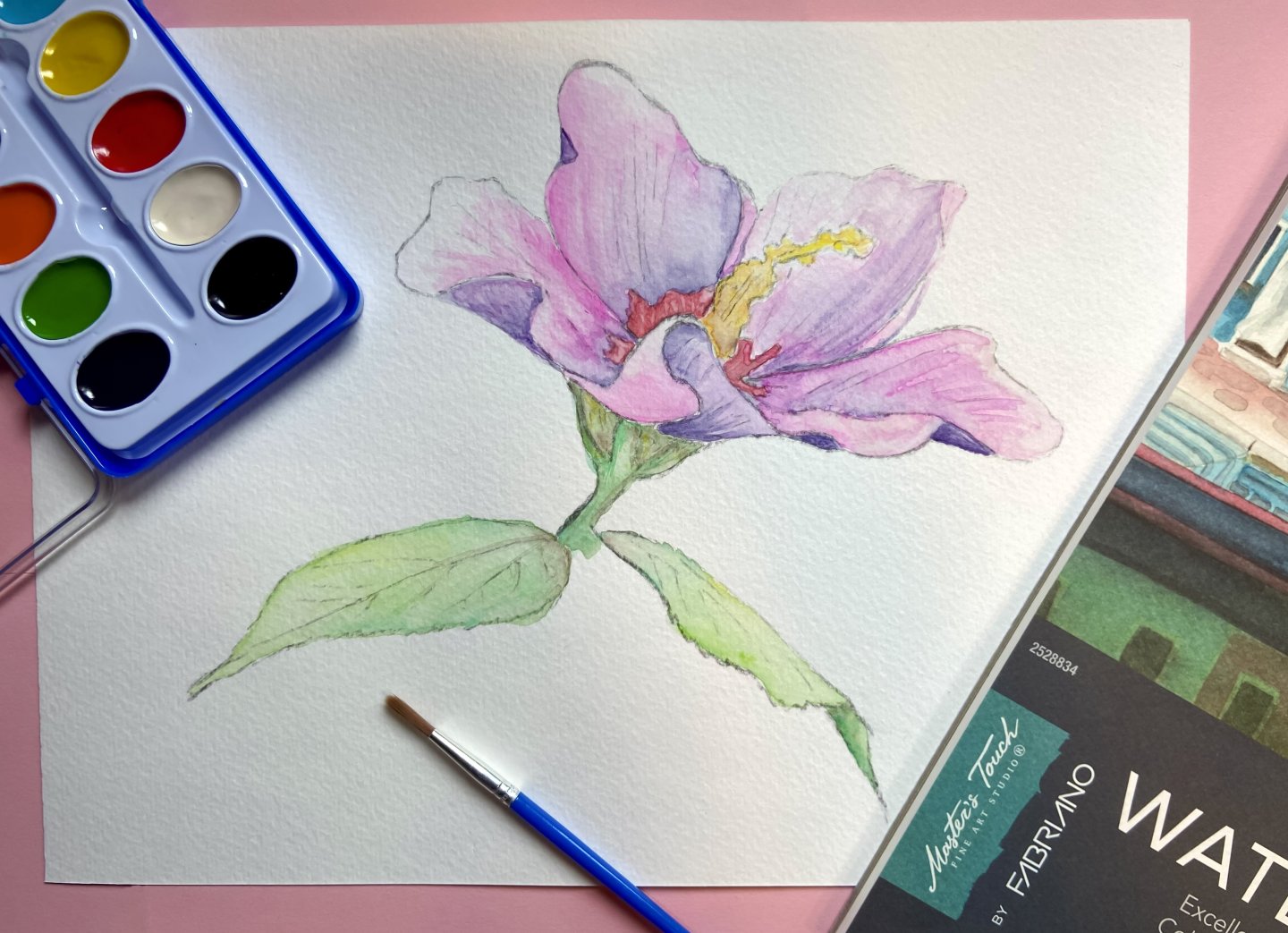 Watercolor painting of a pink flower with green leaves on paper, palette and brush nearby.